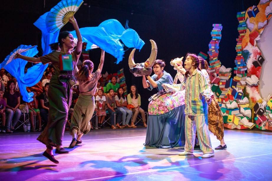 A dancer on stage with large, wing-like woven pieces on their back, in mid-performance.